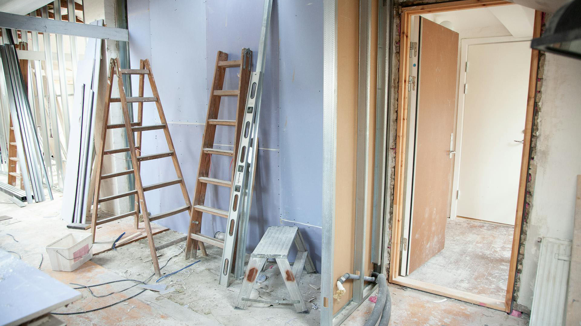 Construction site with wooden ladders, a step stool, and unfinished walls. A doorway leads to another room in a dusty environment.