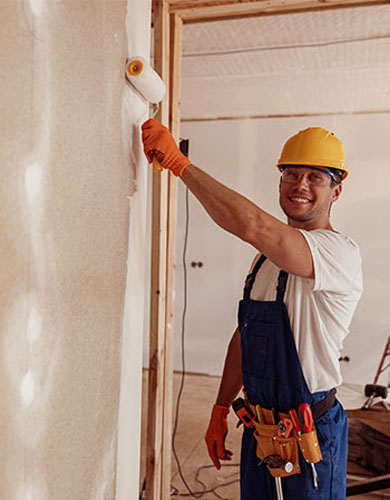 A worker in a yellow hard hat and gloves is painting a wall with a roller, wearing blue overalls and a tool belt.