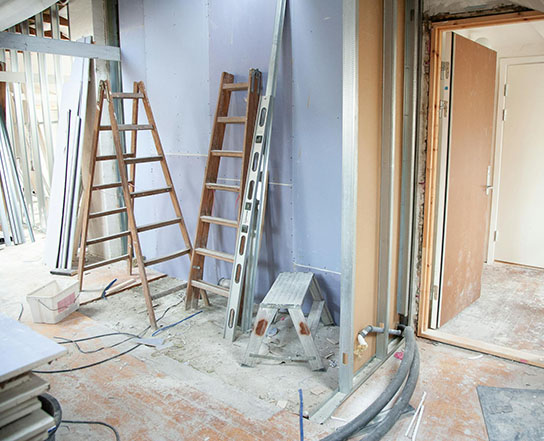 Construction site with wooden ladders, a step stool, and unfinished walls. A doorway leads to another room in a dusty environment.