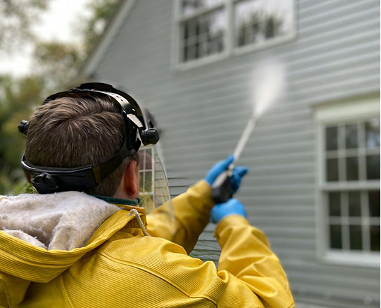 A person in a yellow jacket and gloves uses a power washer to clean the exterior of a house, wearing protective gear.